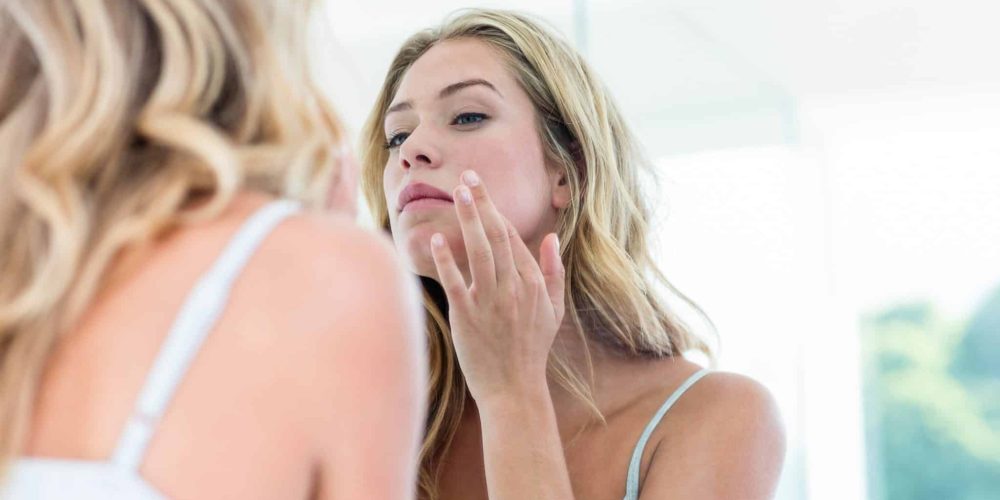 A Young Blonde Woman Cranes Her Head & Examines Her Skin In The Mirror