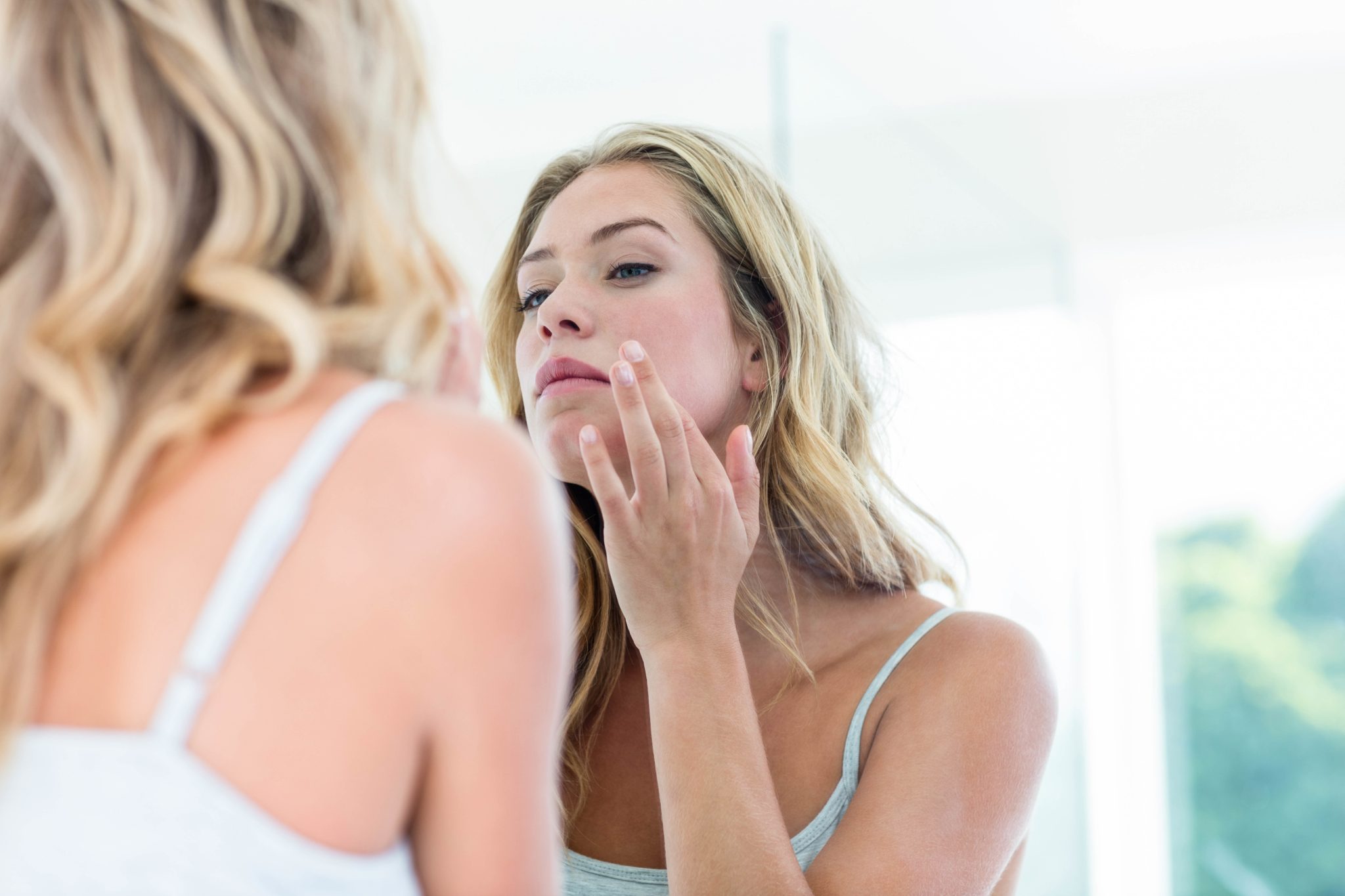 A young blonde woman cranes her head & examines her skin in the mirror