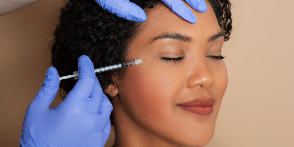 A Woman With Dark Hair Smiles As She Receives Dermal Filler To Smooth Out The Appearance Of Fine Lines And Wrinkles Around Her Eyes.