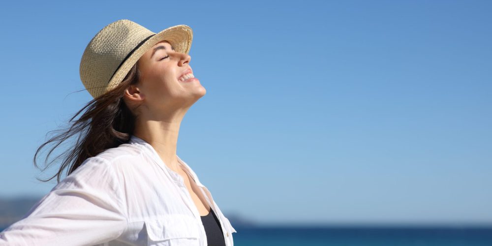 A Woman In A Hat And White Shirt Stands On The Beach, Demonstrating Sun Protection For Skin Health.