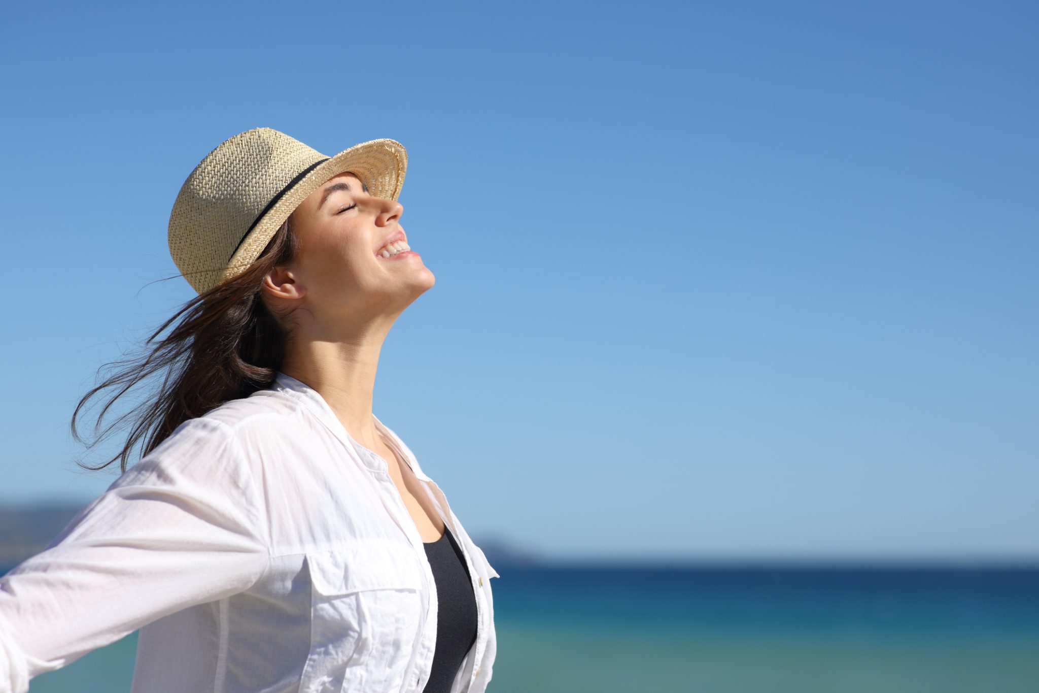 A woman in a hat and white shirt stands on the beach, demonstrating sun protection for skin health.
